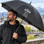 Parapluie noir personnalisable avec logo, photo ou texte, tenu par un homme en extérieur sous la pluie.