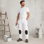 Peintre en bâtiment portant un pantalon ilkott de travail blanc renforcé avec genouillères noires dans une ambiance chantier rénovation BTP.