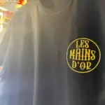 Gros plan du logo poitrine Les Mains d’Or imprimé sur un t-shirt noir suspendu dans l’atelier Pilou Shop 64