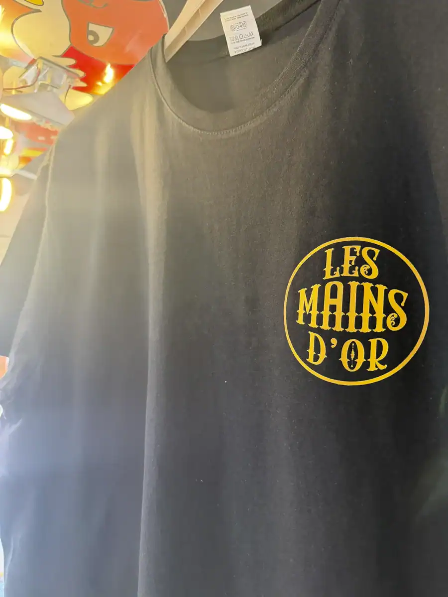 Gros plan du logo poitrine Les Mains d’Or imprimé sur un t-shirt noir suspendu dans l’atelier Pilou Shop 64