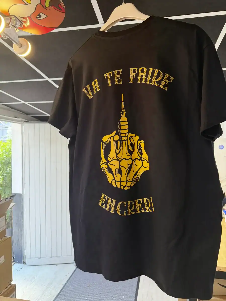 Gros plan du dos du t-shirt Les Mains d’Or avec le design Va te faire encrer suspendu dans l’atelier Pilou Shop 64